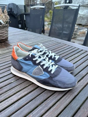 Blå och grå sneakers från Philippe Model - Snygga sneakers från Philippe Model i blått och grått med detaljer i orange vid hälen. Skorna har en mix av mocka och mesh, vita skosnören och en vit sula. Klassisk låg modell med logotyp på sidan. Perfekta för en trendig streetwear-look.