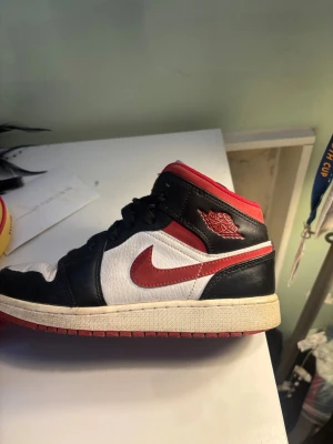 Air Jordan 1 Mid svart/röd/vit - Säljer ett par Air Jordan 1 Mid sneakers i svart, vitt och rött. Skorna har klassisk röd Swoosh, svart läderdetaljer och vit textil på ovandelen. Röd yttersula och vadderad krage för extra komfort. Skorna är i bra skick💞