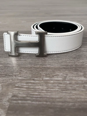 Vit skinnbälte med H-spänne - Säljer ett stilrent vitt skinnbälte från Her”m”s med ett stort metallspänne i form av bokstaven H. Bältet har en slät yta och kontrastsömmar längs kanterna. Perfekt för att lyfta din outfit med en exklusiv touch. 