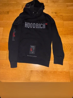 Svart hoodie från Hoodrich - Svart hoodie från Hoodrich med stor logga på bröstet och detaljer i rött och vitt. Har huva, magficka och tryck på både ärm och nederkant. Perfekt för en streetwear-look och riktigt skön passform.