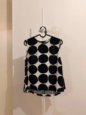 Svartvit prickig linnetopp Marimekko - Säljer en stilren ärmlös topp från Marimekko med stora svarta prickar på vit botten. Toppen har rund halsringning och rak passform, perfekt för en trendig och grafisk look. Materialet känns som bomull och toppen är lätt och luftig.