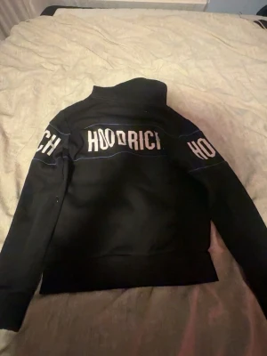 Svart hoodie från Hoodrich - Säljer en svart hoodie från Hoodrich med stor vit logga över bröstet och armarna. Hoodien har huva med dragsko och ribbade muddar vid ärmslut och midja. Perfekt för dig som gillar streetwear och vill ha en snygg och bekväm stil.