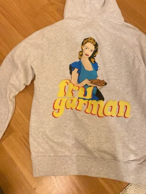 Grå hoodie Fru Garman från Revanite - Cool grå hoodie från Revanite med stor färgglad tryck på ryggen av en retro kvinna som håller en paj och texten 'Fru Garman' i gul och röd. Mjuk bomullsmix och klassisk huva med dragsko. Perfekt för dig som gillar unika prints och retrostil.