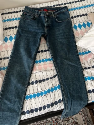 Blå jeans från Hugo Boss - Snygga blå jeans från Hugo Boss med klassisk femficksdesign och rakt ben. Jeansen har en diskret tvättad look och röd Hugo Boss-logga på bakfickan. Perfekta för en stilren och avslappnad outfit. Passar nån som är 165 fast benen är lite långa
