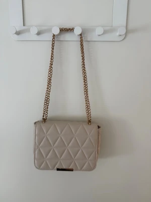 Beige quiltad axelväska med kedja - Snygg beige axelväska med rutigt   mönster och guldfärgad kedjerem.