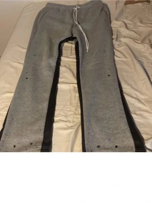 Grå mjukisbyxor med svarta detaljer - Säljer ett par grå mjukisbyxor med svarta ränder längs sidorna och färgstänk som ger en unik look. Byxorna har dragsko i midjan för skön passform och är tillverkade i mjukt material, perfekta för chill dagar.