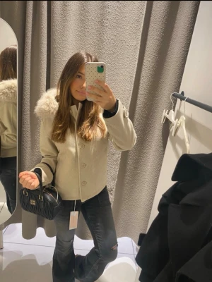 Beige kappa med fuskpäls - Stilren beige kappa med stor krage i fuskpäls och knäppning framtill. Kortare modell som ger en trendig look. Perfekt för dig som vill ha en snygg och mysig jacka till hösten eller vintern. Matcha med jeans och boots för en cool outfit.