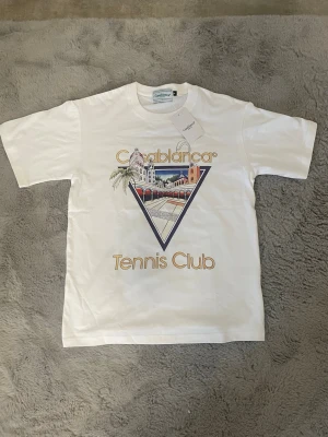 Vit Casablanca Tennis Club t-shirt - Snygg vit t-shirt från Casablanca med färgglatt tryck på bröstet där det står 'Casablanca Tennis Club' och ett grafiskt motiv. Klassisk passform och rund hals. Perfekt för dig som gillar stilrena plagg med coola prints.