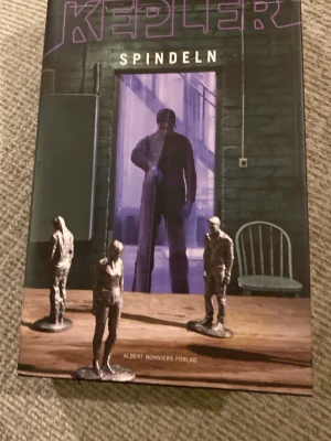 Spindeln av Kepler - En spännande kriminalroman med mörkt omslag där en mystisk figur syns i lila sken bakom en dörr. Omslaget har även små statyetter på ett träbord och en stol i bakgrunden. Perfekt för dig som gillar thrillers och deckare.