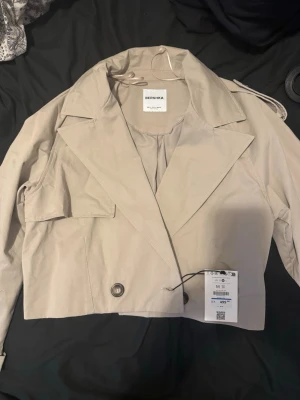 Kort beige trenchcoat från Bershka - Trendig kort trenchcoat från Bershka i beige. Oanvänd då den inte kom till nytta! Pris kan alltid diskuteras💕