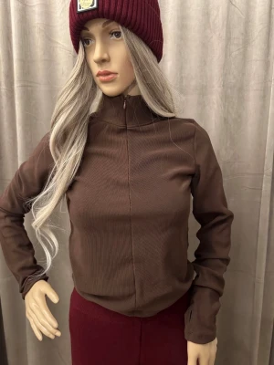 Brun ribbad polotopp med dragkedja XS-S - Snygg brun långärmad polotopp med ribbad struktur och halv dragkedja framtill. Perfekt för lager-på-lager och enkel att styla till olika outfits. Toppens material känns mjukt och följsamt mot kroppen.