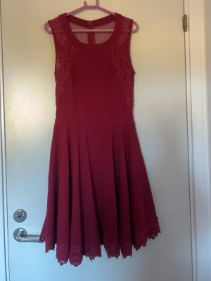 Röd ärmlös klänning med spetsdetaljer - Snygg röd klänning med ärmlös design och rund halsringning. Klänningen har spetsdetaljer vid axlar och nederkant samt meshpartier upptill. Kjolen är utställd och slutar ovanför knät. Perfekt för dig som vill ha en feminin och trendig look.