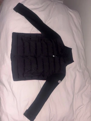 Svart  Moncler cardigan - Snygg svart från Moncler med quiltad front och stickade ärmar. Hög krage och dragkedja framtill. Klassisk Moncler-logga på ena ärmen. Perfekt för lager-på-lager och streetstyle.