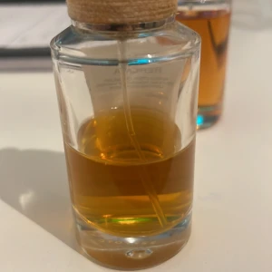 Transparent parfymflaska med träkork - Stilren parfymflaska i genomskinligt glas med träkork och doftsticka. Flaskan har en rund form och innehåller en bärnstensfärgad vätska. Perfekt som inredningsdetalj och för att sprida en härlig doft i rummet.