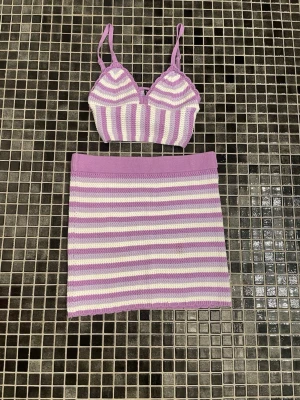 Lila och vitrandigt stickat set - Säljer ett stickat set med en crop top och kjol i lila och vitt. Toppen har smala axelband och V-ringning, medan kjolen är rak och midjekort. Setet är gjort i mjukt garn och har ett randigt mönster. Perfekt för dig som gillar färgglada och unika plagg.