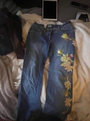 Blå jeans med blommigt tryck - Unika blå jeans med färgglada blommiga tryck längs ena benet och baktill, Från märket philosophy di alberta Ferretti. Jeansen har raka ben, klassisk femficksmodell och bälteshällor. Perfekta för dig som vill sticka ut med en kreativ och personlig stil. Bra kvalitet.( hade sååå använt om de va i min storlek)
