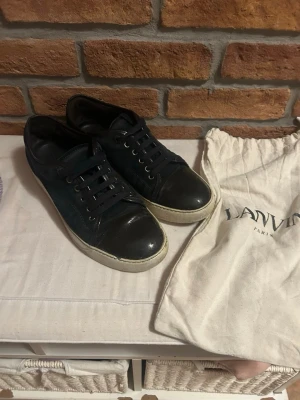 Svarta Lanvin sneakers i läder - Snygga svarta sneakers från Lanvin med ovandel i läder och mocka, vita gummisulor och matchande svarta skosnören. Klassisk låg modell med diskret logga på sulan. Medföljer original dustbag från Lanvin.