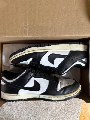 Nike Dunk Low svart/vit sneaker - Klassiska Nike Dunk Low sneakers i svart och vitt läder med svart Swoosh och vita paneler. Skorna har svart yttersula och vita detaljer, samt låga snörade skaft. Perfekta för dig som gillar retrostil och streetwear.