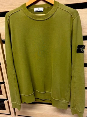 Grön sweatshirt från Stone Island - Säljer en grön sweatshirt från Stone Island med klassisk rund hals och ribbade muddar. Tröjan har den ikoniska Stone Island-loggan på vänster ärm och är tillverkad i mjuk bomull. Perfekt för en avslappnad och stilren look.
