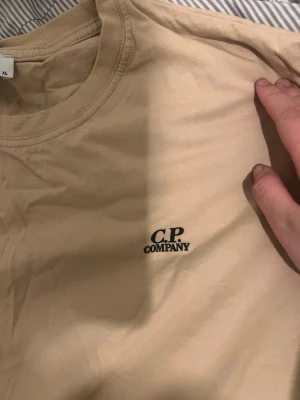 Beige t-shirt från C.P. Company - Snygg beige t-shirt från C.P. Company med diskret logga på bröstet. Klassisk rund halsringning och korta ärmar. Tillverkad i mjuk bomull som känns skön mot huden. Perfekt till jeans eller shorts för en clean och enkel stil.