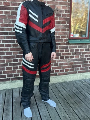 Svart och röd motorcykelställ i skinn - Säljer ett svart motorcykelställ i skinn med röda och vita detaljer. Jackan har dragkedja, bröstficka och markerade axlar. Byxorna har förstärkningar vid knäna och matchande ränder. Snygg retrostil och perfekt för dig som gillar mc-kläder.