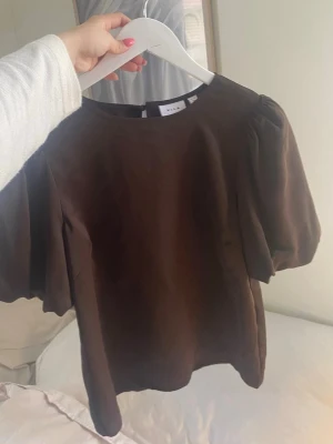 Brun puffärm topp från Vila - Snygg brun topp från Vila med puffiga korta ärmar och rund halsringning. Tillverkad i ett mjukt material som ger en stilren look. Perfekt att matcha med både jeans och kjol för en trendig outfit.