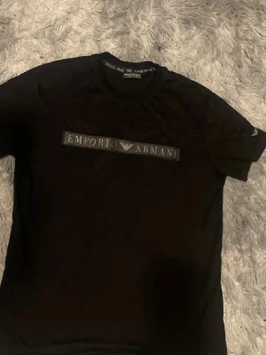 Svart Emporio Armani t-shirt - Snygg svart t-shirt från Emporio Armani med klassisk logga framtill. T-shirten har rund halsringning och korta ärmar. Perfekt för dig som gillar stilrena och exklusiva plagg med en modern touch.