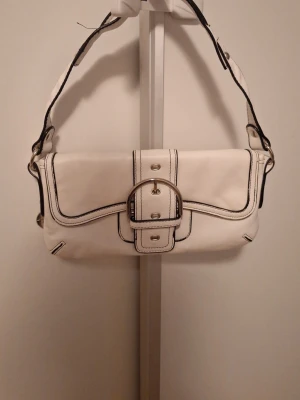 Vit axelväska - Snygg vit axelväska med svart kontrastsöm och stor dekorativ metallspänne framtill. Väskan har ett justerbart axelband och flera innerfack, inklusive dragkedjefack. Fodret är beige med logotypmönster. Perfekt för dig som gillar stilrena detaljer.