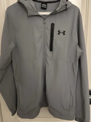 Grå vindjacka från Under Armour - Snygg grå vindjacka från Under Armour med huva och dragkedja framtill. Jackan har en svart logga på bröstet och en praktisk bröstficka med dragkedja. Perfekt för blåsiga dagar och sportiga outfits.