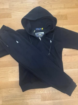 Polo Ralph Lauren Svart tracksuit, Fullzip Hoodie + Byxor - Säljer ett svart Polo Ralph Lauren set med hoodie (dragkedja) och matchande joggerbyxor.  Helt nytt, alla tags sitter kvar.  Storlek M men liten i storleken – sitter mer som S.  Väldigt stilrent och skönt set, säljer då det inte kommit till användning. 