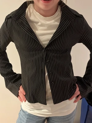 Svart kavaj med vita ränder - Snygg svart kavaj med smala vita kritstrecksränder och hög krage. Figurnära modell med långa ärmar och dold knäppning framtill. Perfekt för dig som gillar en stilren och trendig look.