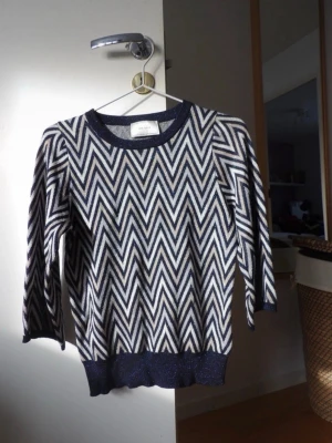 Stickad tröja med zigzag från Neo Noir - Snygg stickad tröja från Neo Noir med zigzag-mönster i marinblått, vitt och beige. Tröjan har rund halsringning, ribbade muddar och en lätt glittrig finish. Står ingen storlek men skulle säga XS-S