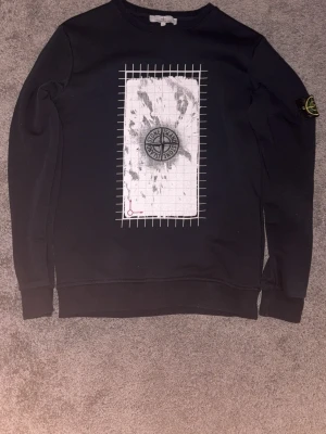 Svart Stone Island sweatshirt M - Svart sweatshirt från Stone Island med tryckt grafiskt motiv och kompasslogga på bröstet. Klassisk rund halsringning och ribbade muddar. Ikonisk logotyp-patch på vänster ärm. Perfekt för en streetwear-look.