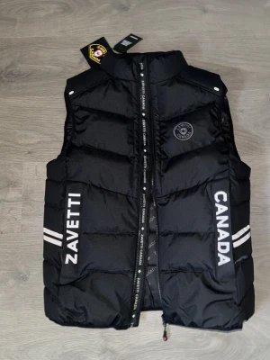 Svart dunväst från Zavetti Canada - Snygg svart dunväst från Zavetti Canada med vita logotexter på framsidan och två vita ränder på sidorna. Västen har hög krage, dragkedja framtill och quiltad design. Perfekt för lager-på-lager och streetwear-stil.