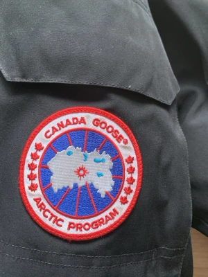 Svart parkas från Canada Goose - Säljer en svart parkasjacka från Canada Goose med det klassiska Arctic Program-märket på ärmen. Jackan har en rak passform och är tillverkad i slitstarkt material som står emot kyla och vind. Perfekt för kalla vinterdagar och riktigt snygg streetstil.