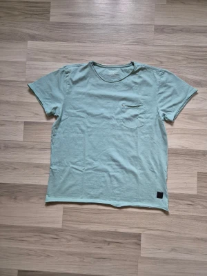 Ljusgrön t-shirt med bröstficka - Snygg ljusgrön t-shirt med rund halsringning och en liten bröstficka med knappdetalj. T-shirten har korta ärmar och en avslappnad passform. Perfekt för en chill och stilren look.
