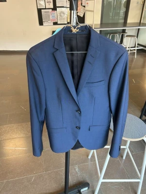 Mörkblå kavaj från Tiger Of Sweden  - Snygg mörkblå kavaj från Tiger Of Sweden med två knappar framtill, klassisk passform och smala slag. Kavajen har tre fickor fram och är helfodrad. Perfekt för dig som vill ha en stilren och tidlös look.