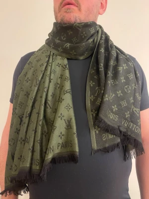 Grön Vuitton halsduk - Stilren grön halsduk från  Vuitton med klassiskt monogrammönster och svarta fransar i kanterna. Halsduken har en lyxig känsla och är tillverkad i ett mjukt tyg som känns skönt mot huden. Perfekt accessoar för att lyfta din outfit.