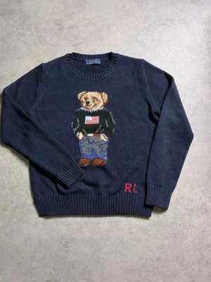 Mörkblå stickad tröja Polo Ralph lauren  - Mörkblå stickad tröja från Polo Ralph Lauren med ikoniska Polo Bear-motivet framtill. Rund halsringning, ribbade muddar och RL-broderi nertill. Hundra procent riktigt med Ralph lauren påsen! Perfekt för dig som gillar klassisk preppy stil och vill ha något som sticker ut. Detta är ett måste!  