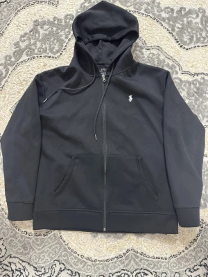 Svart hoodie från Polo Ralph Lauren - Klassisk svart hoodie från Polo Ralph Lauren med dragkedja framtill och vit broderad logga på bröstet. Hoodien har justerbar huva med snören och två fickor framtill. Tillverkad i mjukt bomullsmaterial som är skönt att bära.