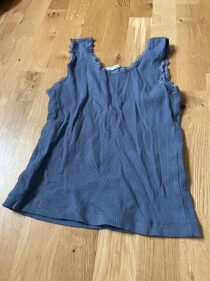 Blå ribbad spetslinne från 157 - Säljer ett blått ribbat linne från 157 med spetsdetaljer vid halsringning och ärmhål. Linnet är ärmlöst och har en mjuk, stretchig känsla som passar perfekt till jeans eller kjol. Enkel och stilren design med en feminin touch.