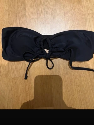 Svart bandeau bikiniöverdel med snörning - Stilren svart bandeau bikiniöverdel med snörning framtill som ger en snygg detalj och möjlighet att justera passformen. Perfekt för soliga dagar på stranden eller vid poolen. Tillverkad i mjukt syntetmaterial som torkar snabbt.