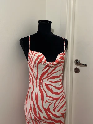 Zebramönstrad långklänning med slits ❣️ - Snygg långklänning med rödvitt zebramönster och tunna axelband. Klänningen har en draperad urringning och en hög slits på sidan för extra attityd. Tillverkad i ett glansigt, mjukt material som ger en lyxig känsla.