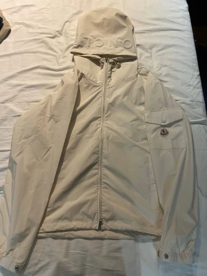 Vit vindjacka från Moncler - Snygg beige vindjacka från Moncler med huva och dragkedja framtill. Jackan har Moncler-logga på ärmen, justerbar huva och elastiska muddar. Tillverkad i lätt och tåligt material som passar perfekt för blåsiga dagar.