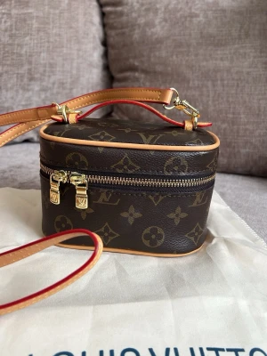 Mini Monogram Vanity Handväska - Snygg brun handväska sminkväska i klassiskt monogrammönster. Väskan har boxig form, guldfärgade detaljer och dragkedja runt om. Ljust beige insida och justerbar axelrem med röda kanter. Perfekt för dig som gillar lyxiga accessoarer.