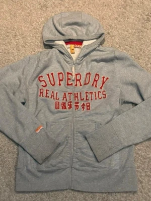 Grå Superdry hoodie med dragkedja - Grå hoodie från Superdry med dragkedja och huva. Mjuk insida och fickor framtill. 