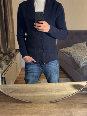 Mörkblå cardigan  - Säljer en mörkblå cardigan med sjalkrage och stora knappar framtill. Cardiganen har två fickor och är ribbstickad för en snygg struktur. Perfekt att slänga över en t-shirt för en avslappnad men stilren stil. Har bara haft på mig den 2 gånger.