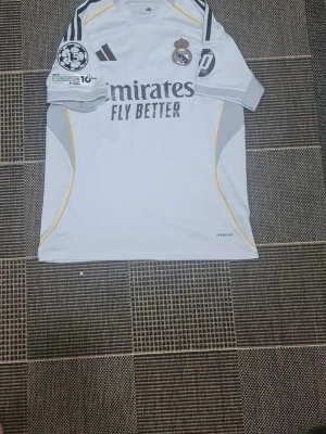 Real Madrid matchtröja Adidas vit - Säljer en vit Real Madrid matchtröja från Adidas med svarta detaljer och gula inslag. Tröjan har klubbmärke, Adidas-logga och 'Emirates Fly Better' tryckt på bröstet. På ärmen finns UEFA Foundation och Champions League-märke. Tillverkad i lätt funktionsmaterial. Jätte fin Mbappe tröja. Inte använd mycket 