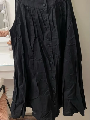 Svart långklänning med knappar - Svart långklänning med lös passform och knappar framtill. Klänningen har ärmlös design och lätt rynkade detaljer vid axlarna för en avslappnad look. Perfekt för dig som gillar enkel och stilren stil.