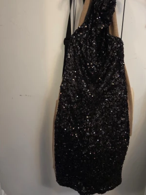 Svart paljettklänning omlott - Säljer en svart kort klänning täckt med glittriga paljetter. Klänningen har omlottdesign och en snygg draperad detalj vid halsen. Perfekt för dig som vill sticka ut och glänsa. Passar dig som gillar trendiga och festliga plagg.